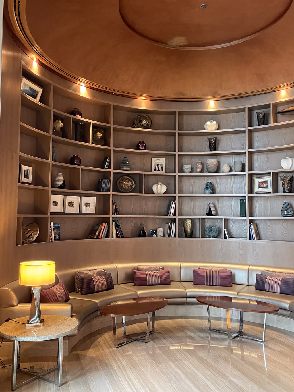 A luxurious, circular seating area with wooden built-in curved bookshelves and ambient lighting in the Andaz Capital Gate hotel.