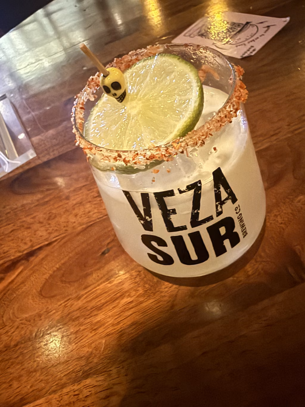 A cocktail in a glass branded with Veza Sur Brewing Co. logo, garnished with a lime and a skull toothpick, sitting on a wooden table.