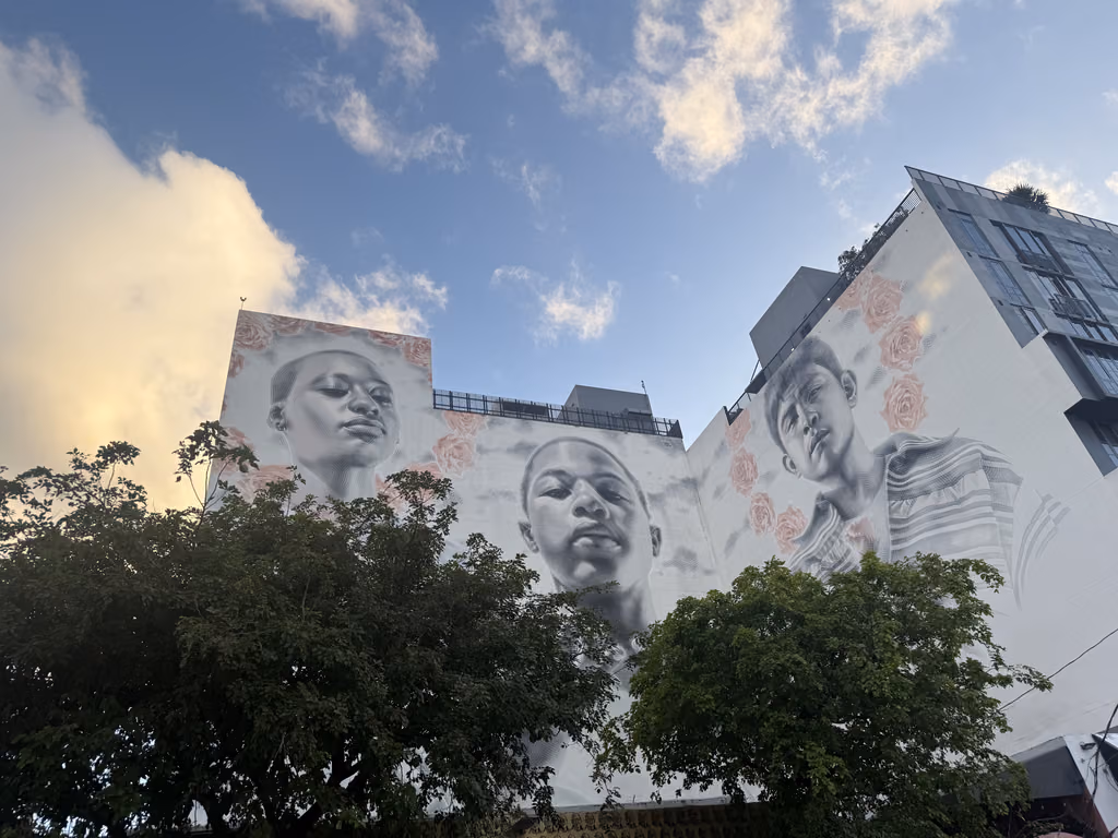 Large-scale monochromatic mural portraits of three people decorated with pink flowers on a Wynwood building facade under a blue sky.