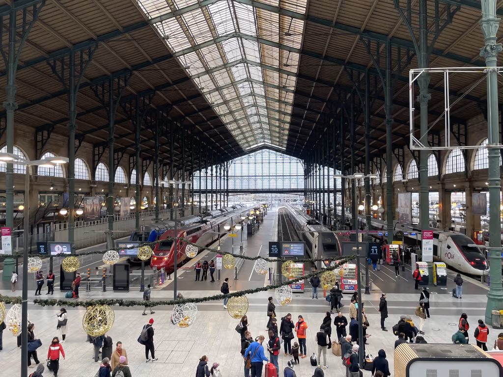 A high-angle view looking down into the bustling, glass-roofed Gare de l'Est railway station in Paris with passengers walking on the platform and trains parked at the tracks.