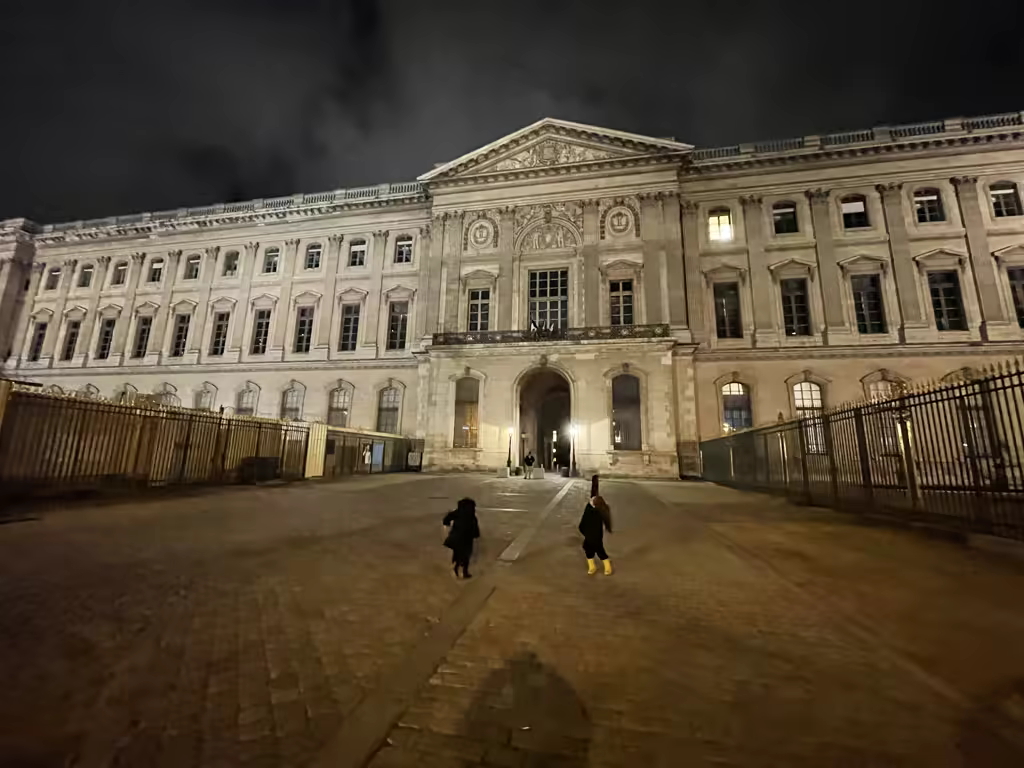 Two children running across the illuminated paved courtyard of the Louvre Museum in Paris at night.