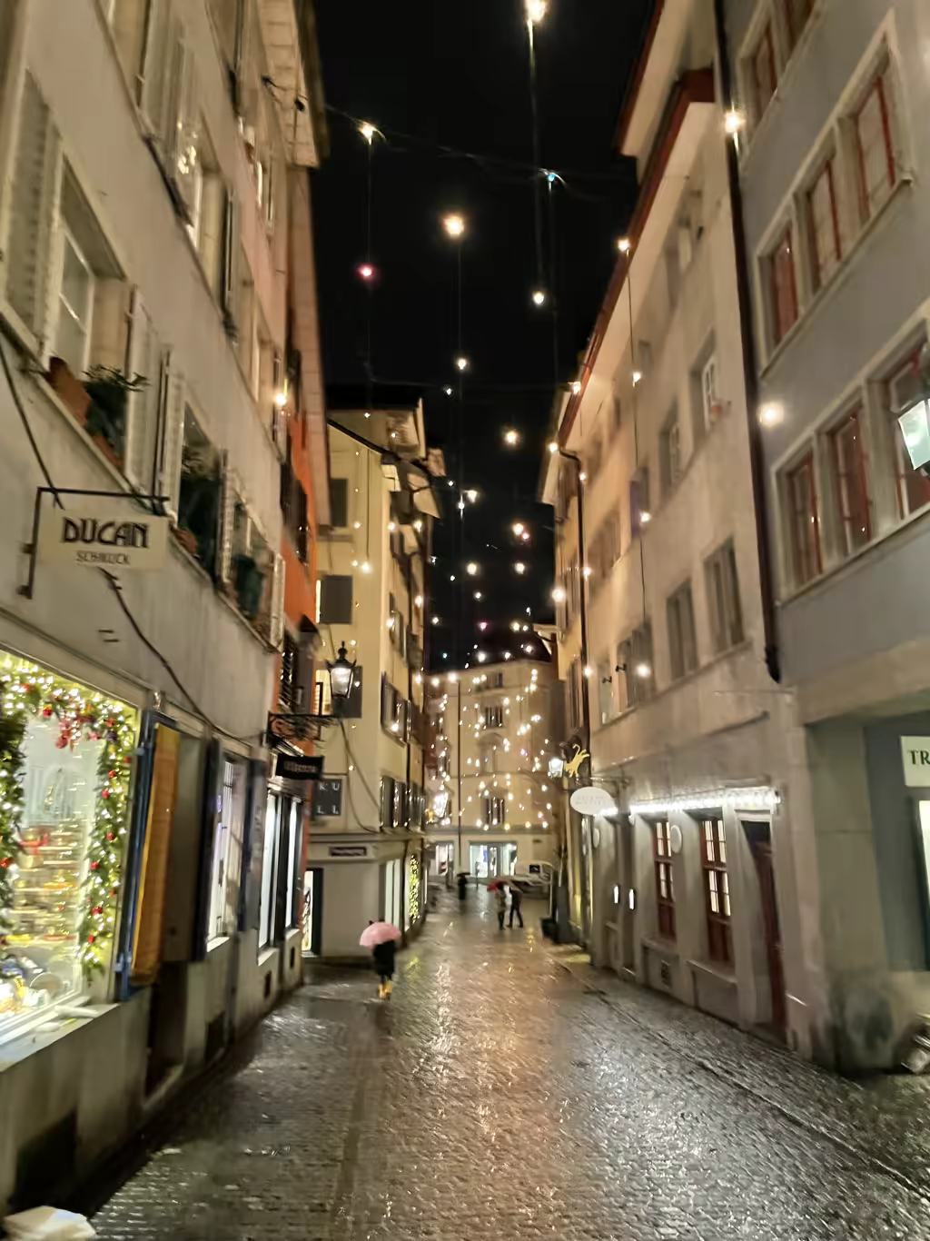 A narrow, rain-slicked cobblestone street in Zurich's Old Town at night, decorated with hanging festive lights during the Christmas season.
