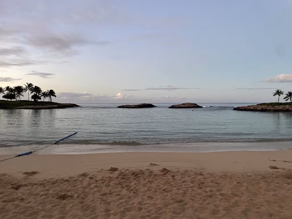 A serene view of a calm, blue lagoon at Disney’s Aulani Resort in Ko Olina, Hawaii, surrounded by sandy shores and palm trees.