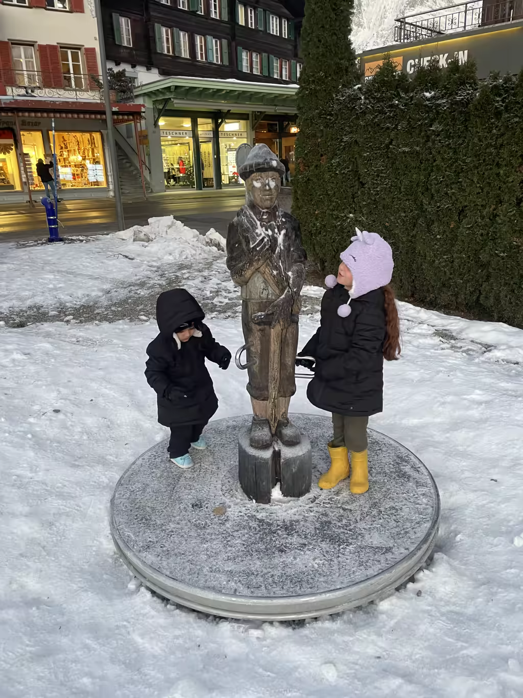 The kids explore a whimsical wooden statue on a snowy street in central Spiez, Switzerland.
