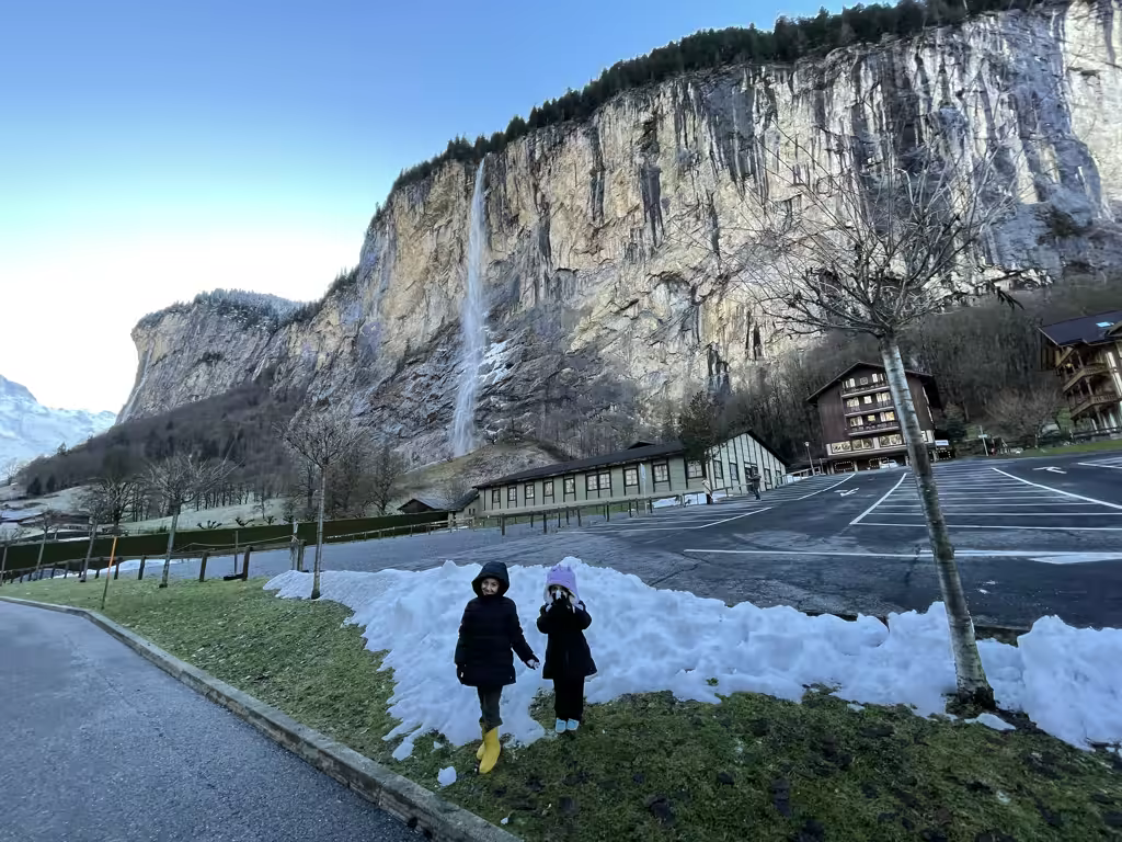 Two children standing by a snowy bank in front of Staubbach Falls, Lauterbrunnen, Switzerland.
