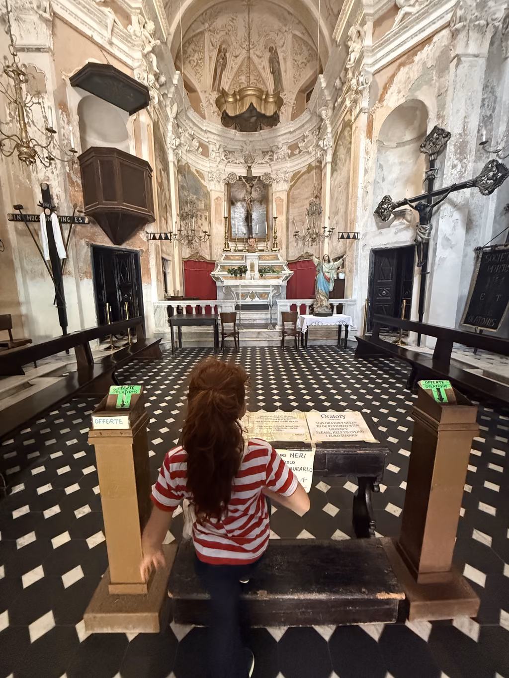 A young girl with long reddish-brown hair in a striped shirt stands in an old Italian Oratory, looking toward the altar.