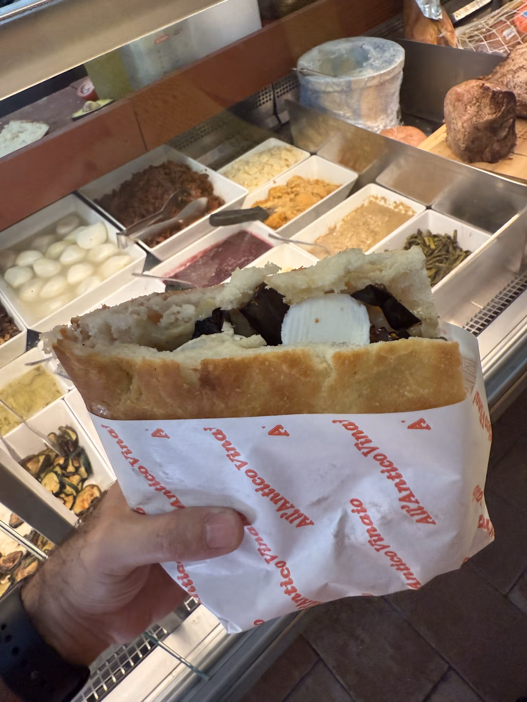 Hand holding a fresh sandwich wrapped in All'Antico Vinaio paper inside a Florence deli.