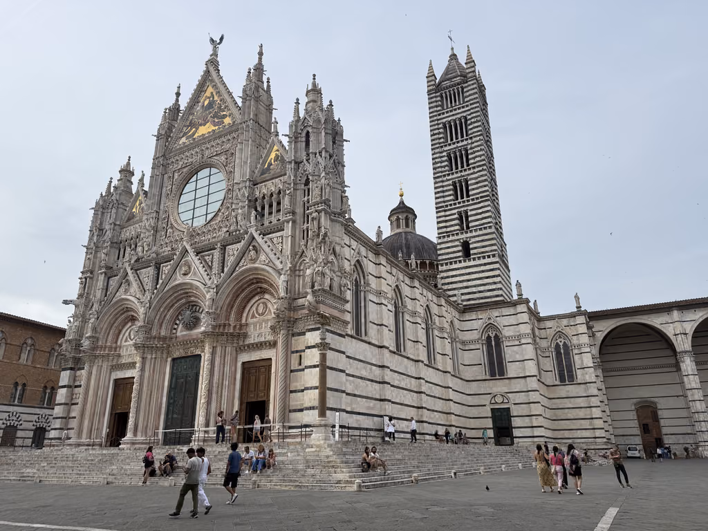 Tourists walk past the grand, striped marble facade and bell tower of the Siena Cathedral in Italy.