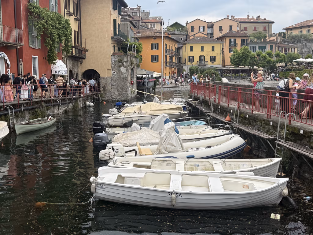 Boats moored at a narrow canal in Varenna, Lake Como, with pedestrians walking along the waterfront.