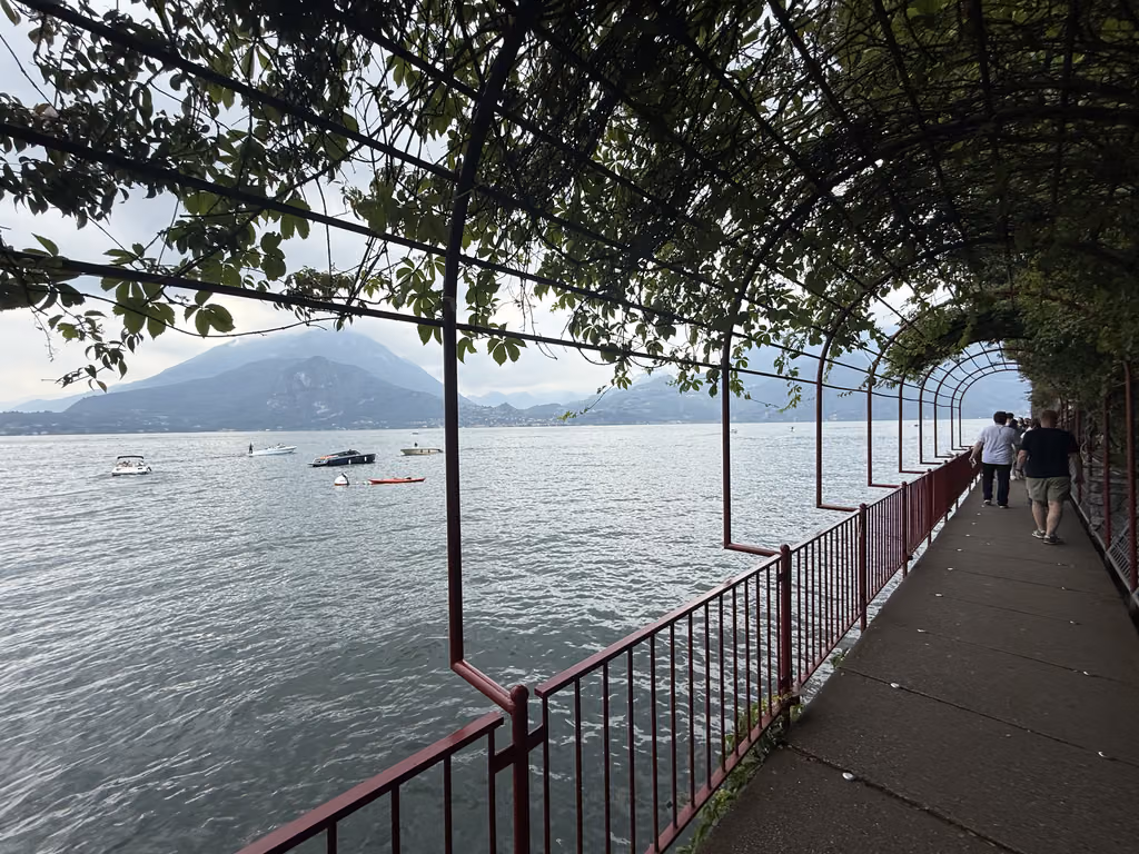 People walking along a leafy, vine-covered waterfront path in Varenna, Lake Como, Italy.