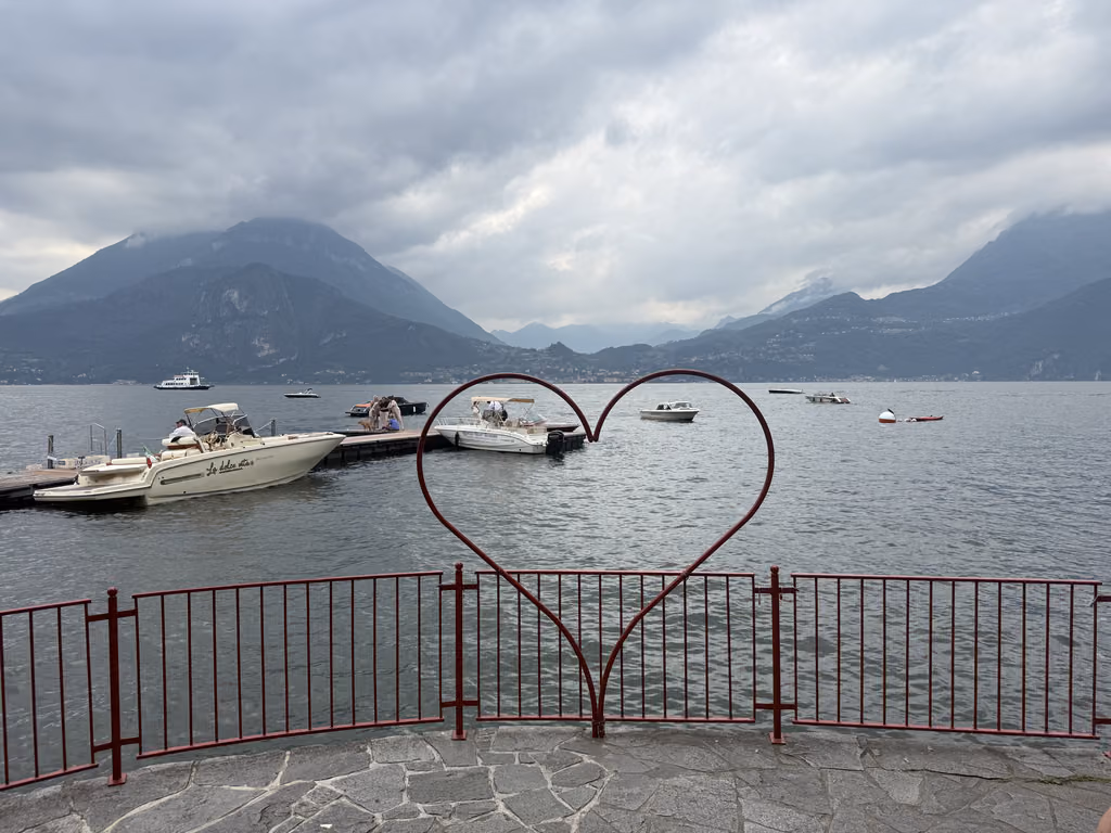 A red heart-shaped sculpture on the Varenna waterfront overlooking Lake Como with boats docked at a pier.