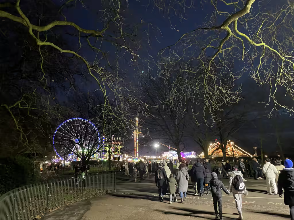Visitors walking through Hyde Park Winter Wonderland at night with an illuminated Ferris wheel and carnival rides in the background.