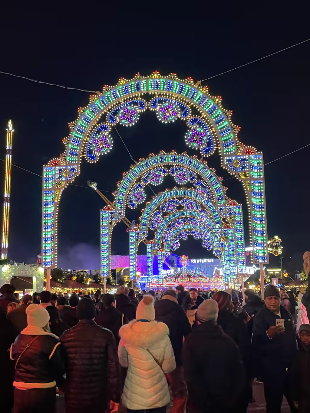 A large crowd of people walking through a series of brightly lit, colorful LED arches at the Winter Wonderland fair in Hyde Park at night.