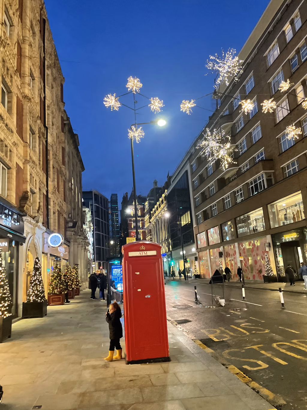 A young traveler stops by a classic red phone booth during a festive evening walk.