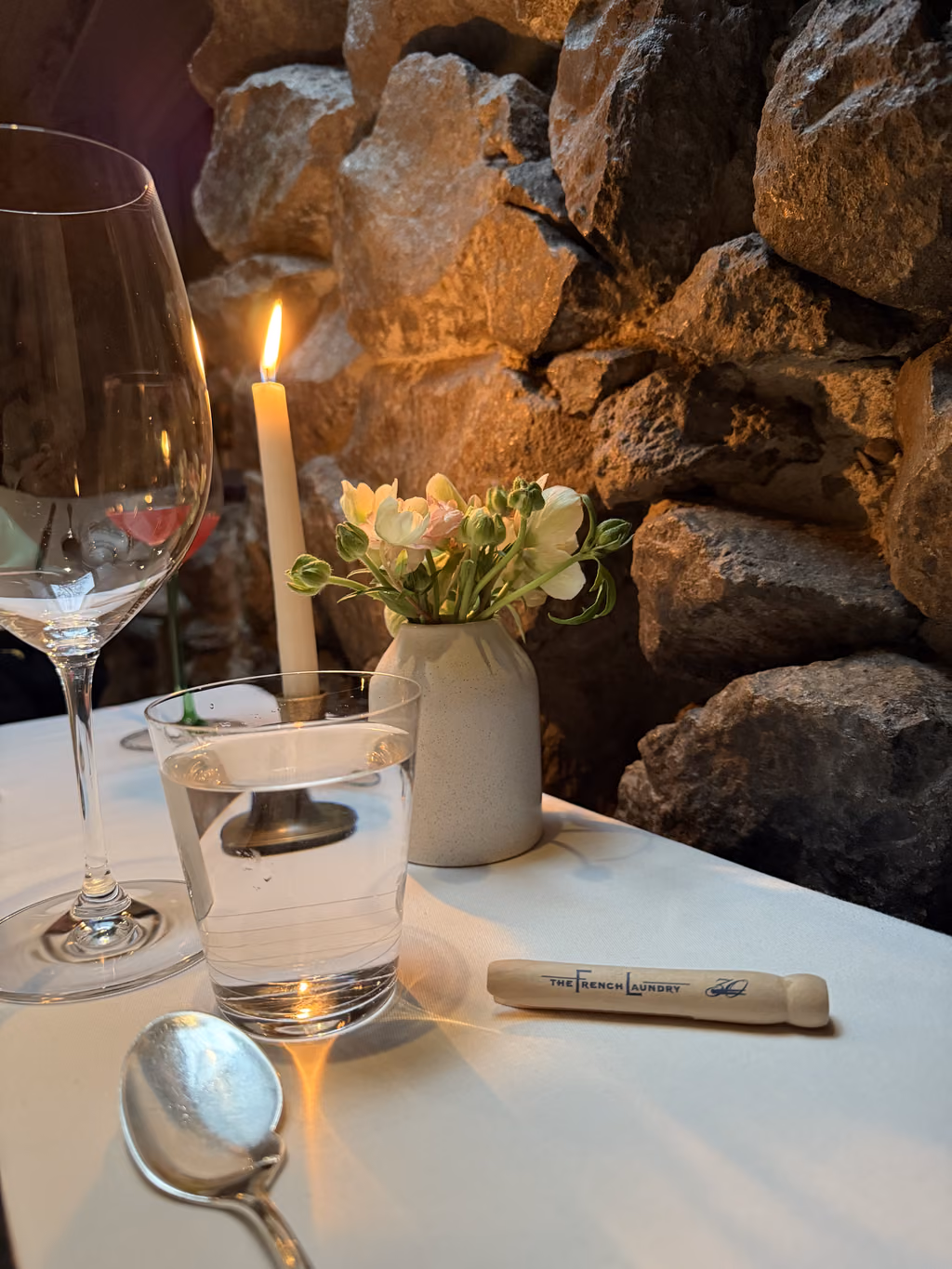 A candlelit table setting at The French Laundry featuring a wine glass, a water tumbler, a small flower vase, and a wooden souvenir clothespin.