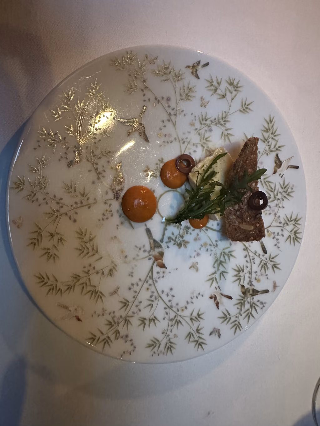 A gourmet amuse-bouche appetizer featuring a slice of cheese, rye bread, and olive served on a decorative plate at The French Laundry in Napa.