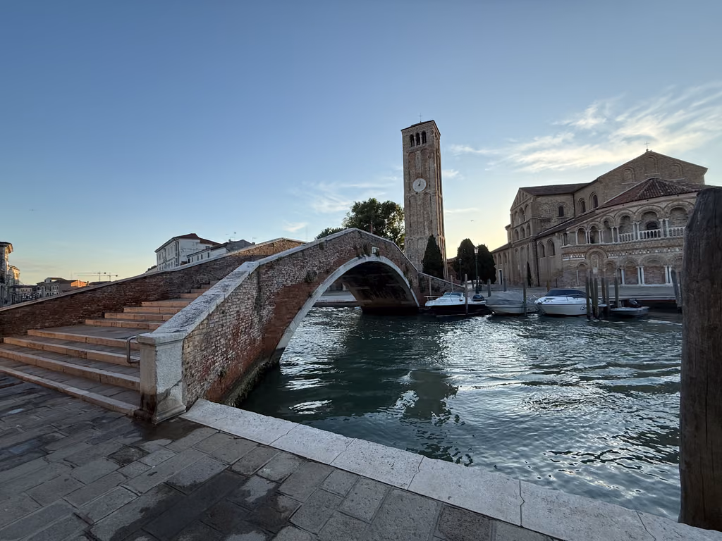 A scenic view of the red brick bridge and the historic bell tower of the Basilica di Santa Maria e San Donato in Murano, Venice, with boats docked in the canal at sunset.