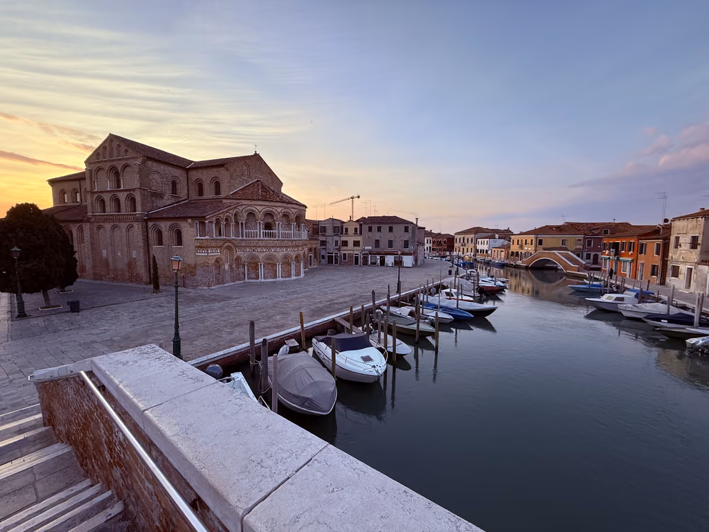 Sunset view over a canal in Murano, Venice, featuring the historic Basilica di Santa Maria e San Donato and moored boats.