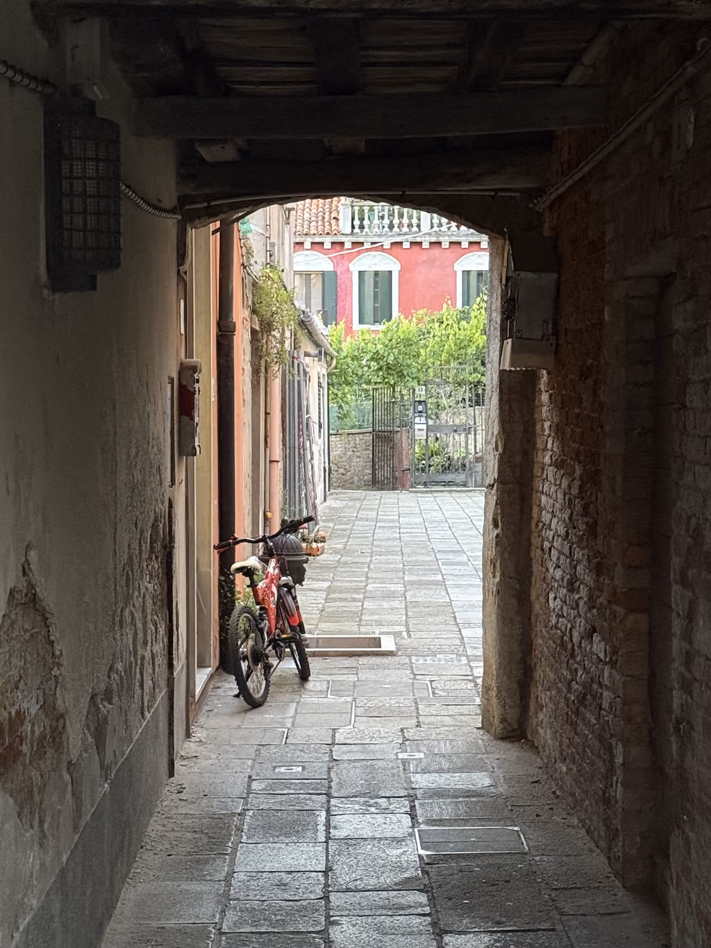 A narrow stone alleyway in Murano, Venice, featuring a parked bicycle and a red building in the distance.