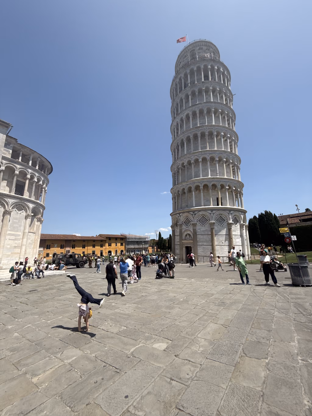 Child doing a handstand in the square in front of the Leaning Tower of Pisa under a clear blue sky.
