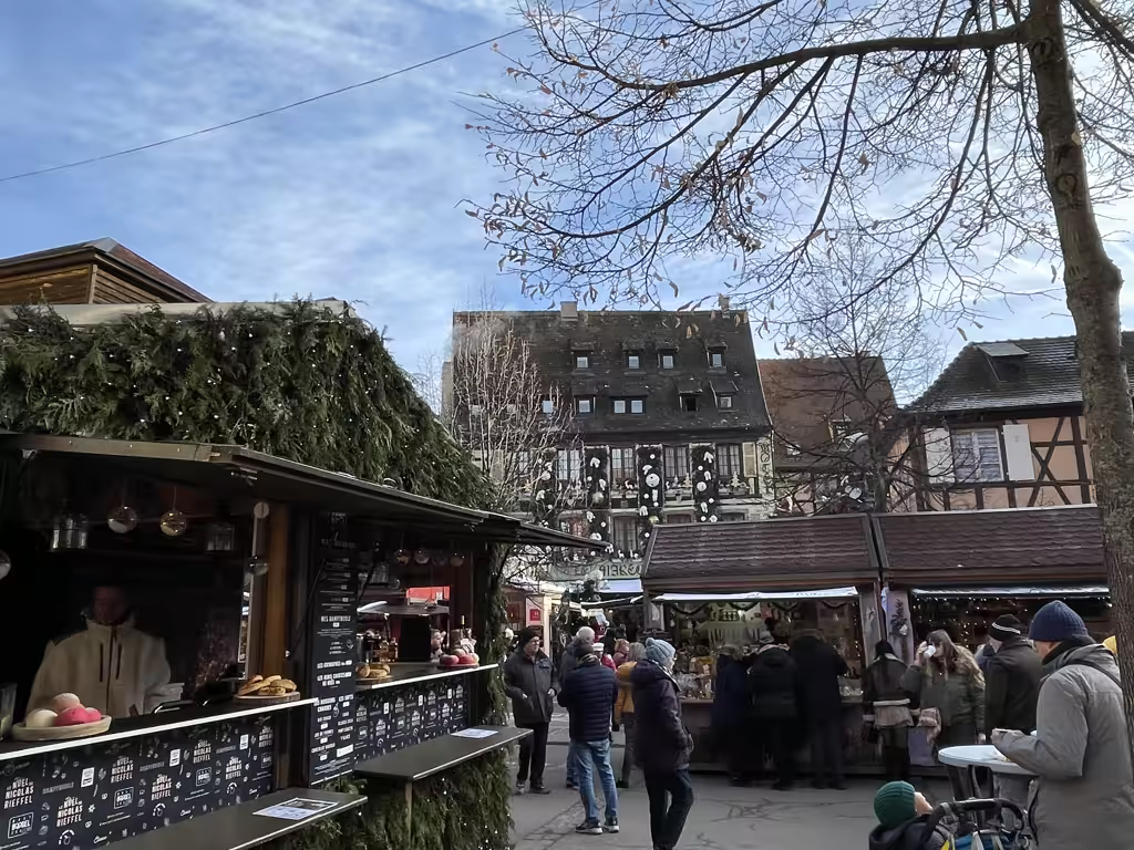 Crowded Christmas market stalls in Colmar, France, featuring timber-framed buildings in background.
