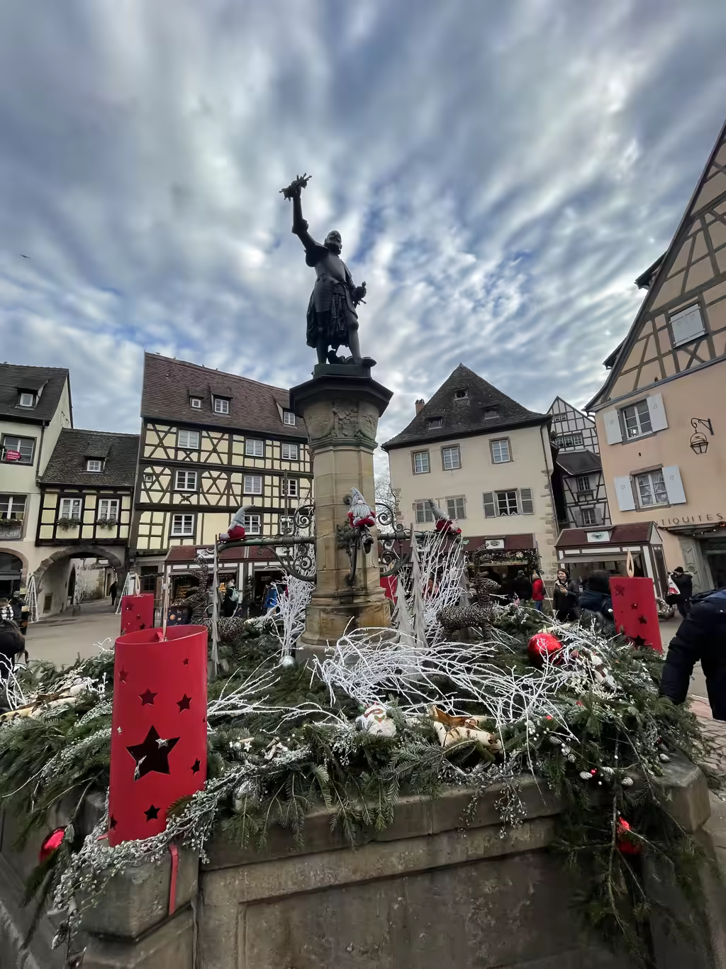 Charming half-timbered buildings surround the historic Roesselmann Fountain during a festive winter afternoon in Colmar.