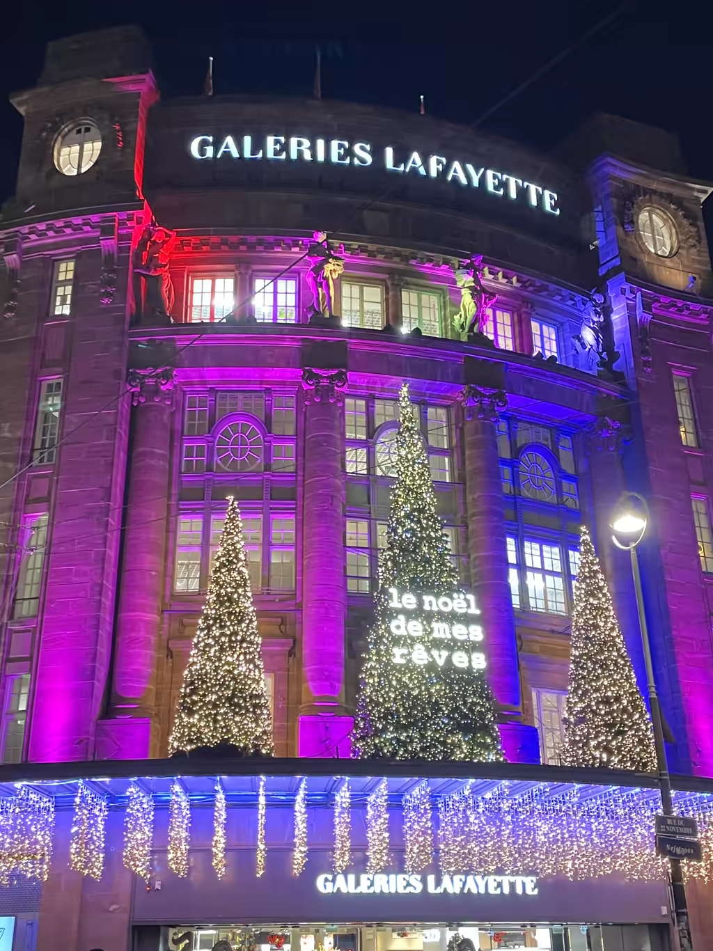 The festive facade of Galeries Lafayette in Strasbourg sparkles with stunning purple lights and Christmas trees.