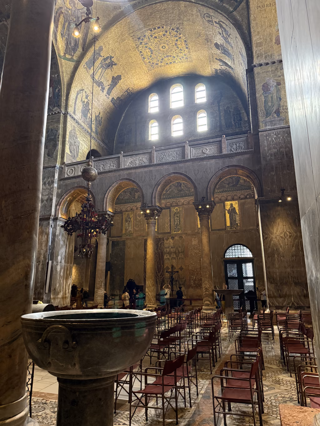 Interior view of St. Mark's Basilica featuring a large stone baptismal font and golden mosaics.